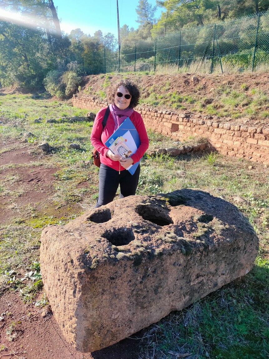 Tamara Lewit stands behind a large Roman weightstone in a field.