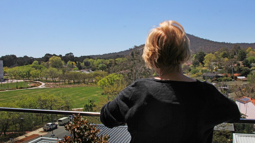 Lady wearing a black shirt overlooks the balcony at a field surrounded by trees and older looking houses.