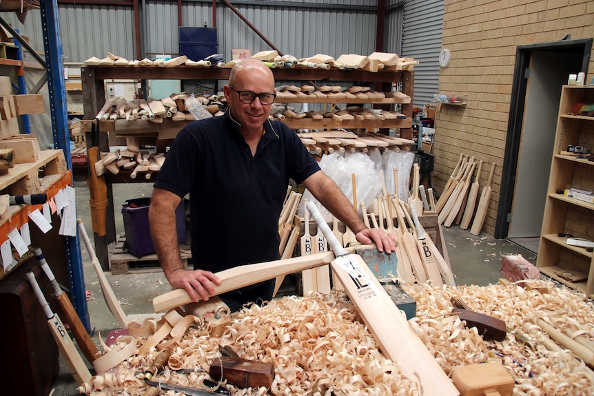 Paul stands holding an unfinished cricket bat in front of a pile of woodshavings.