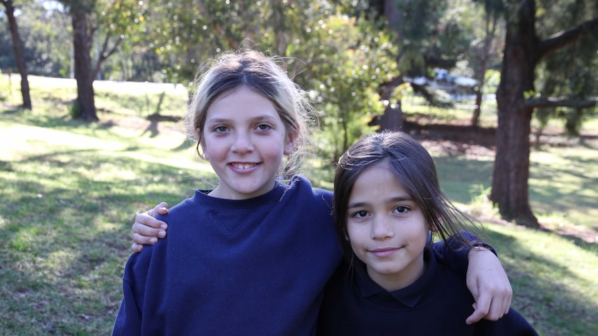 Two girls, left is taller, standing with arms around each others shoulders, background is out of focus greenery.