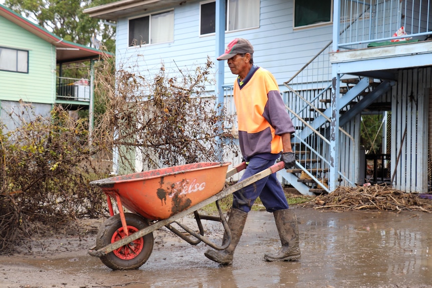 A man in work gear pushes a wheelbarrow past a flood-affected house.