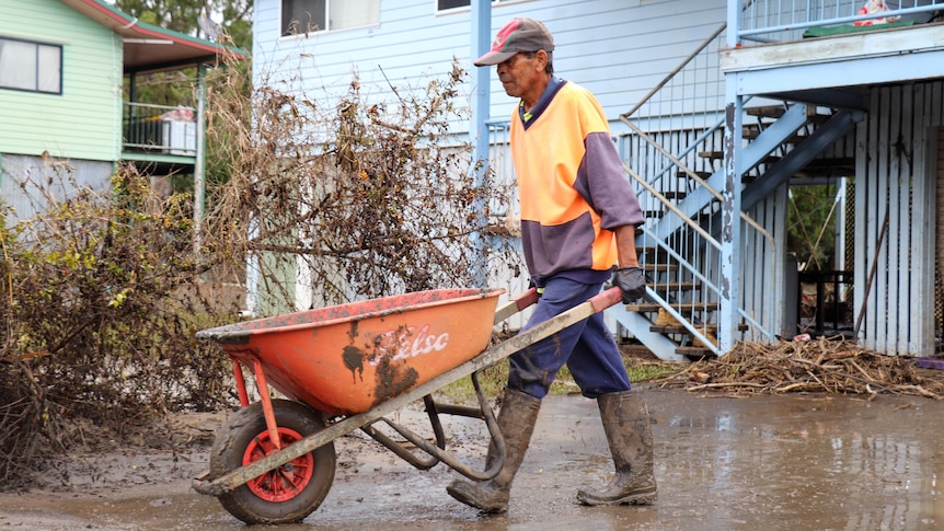 A man in work gear pushes a wheelbarrow past a flood-affected house.