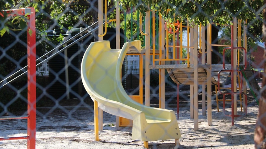 A playground seen through a wire fence featuring a yellow slide and wooden play equipment.