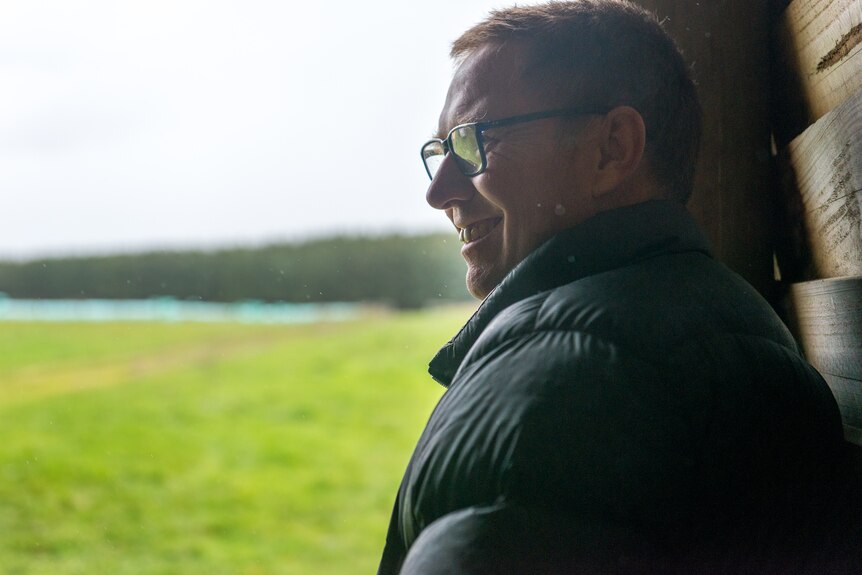 A man leans against a wood paneled wall smiling, grassy paddocks behind him.