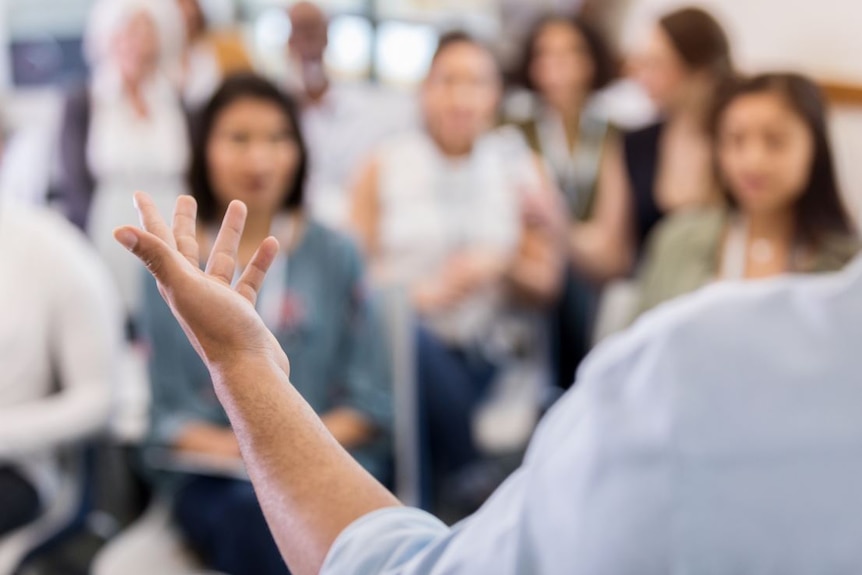 A person - only part of a shoulder with an outstretched arm and hand is visible - stands before a blurred group of people.