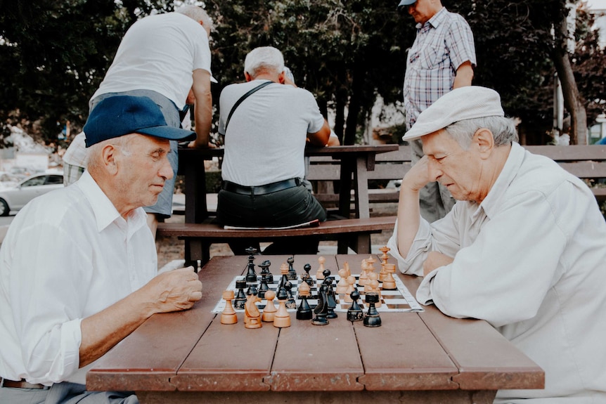 Two older men sitting down and playing chess with other men in the background