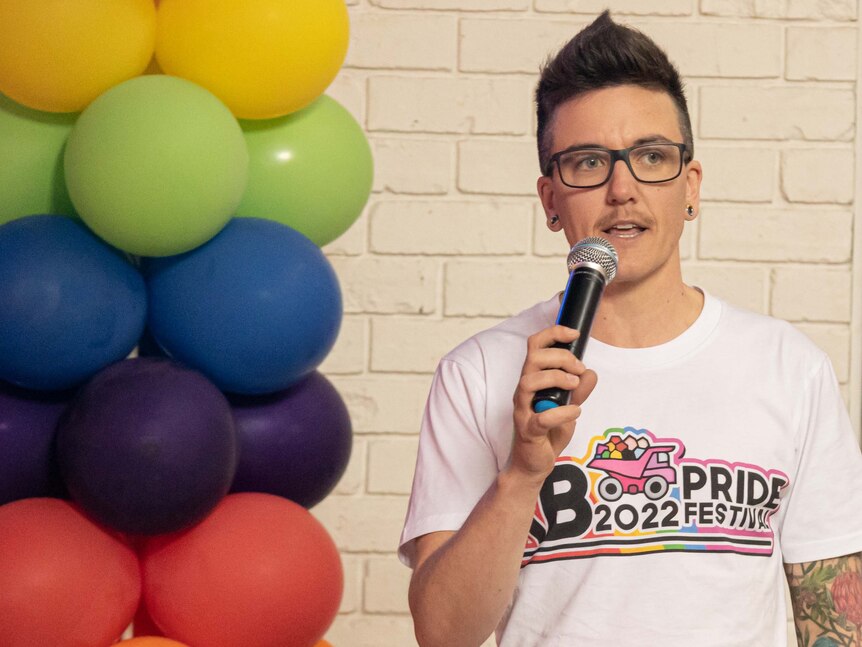 A young person with short hair and glasses wearing a pride t-shirt and holding a mic.
