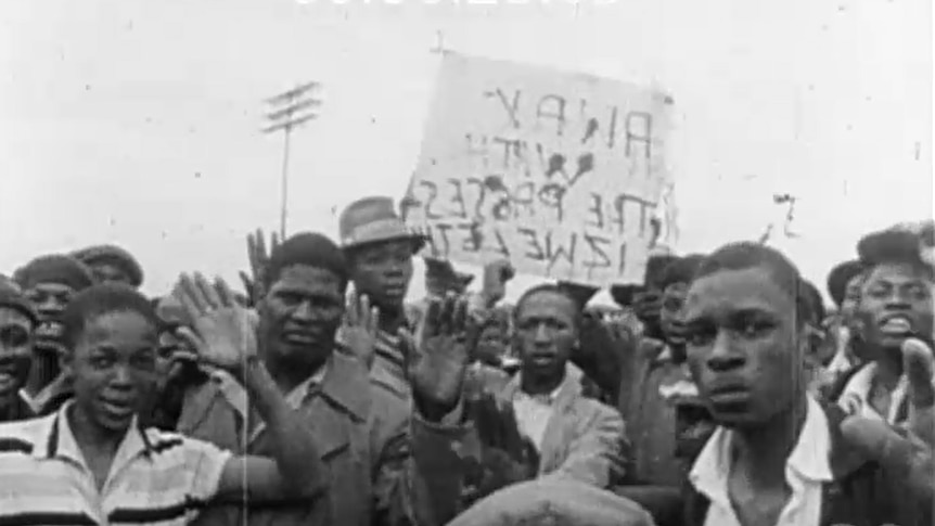 A black and white screenshot of footage showing a group of Black South Africans protesting. 