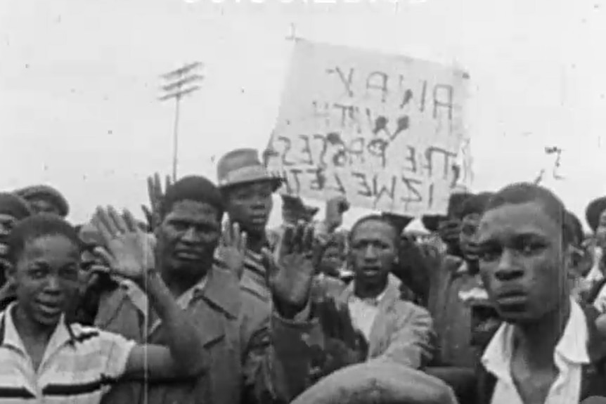 A black and white screenshot of footage showing a group of Black South Africans protesting. 