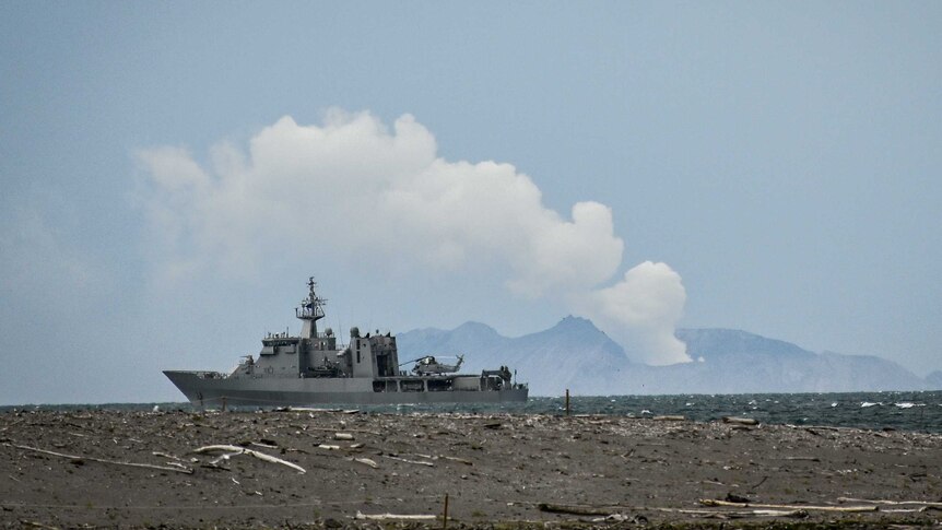 A navy boat in the foreground with a smoke cloud billowing from a volcano in the background