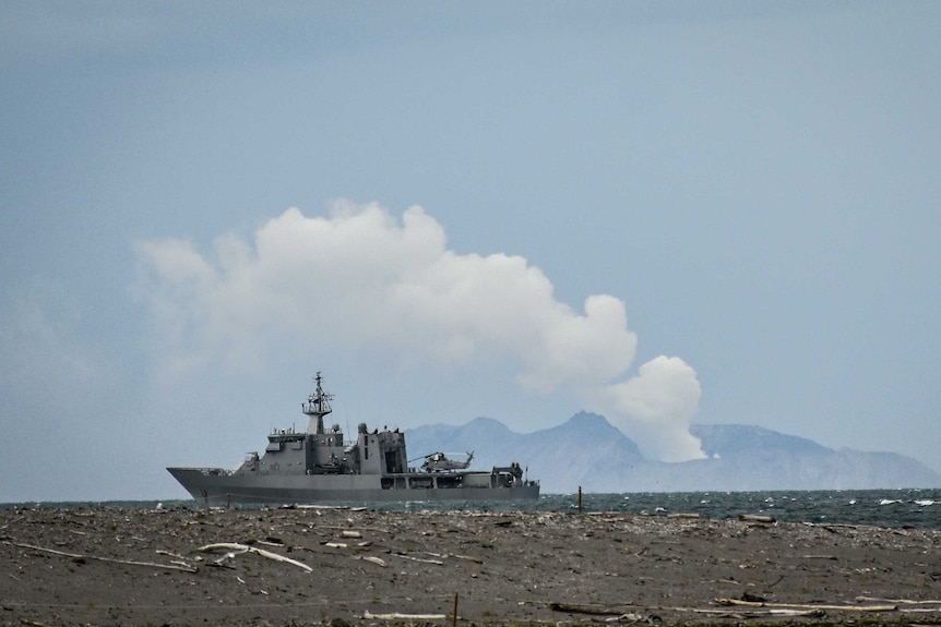 A navy boat in the foreground with a smoke cloud billowing from a volcano in the background