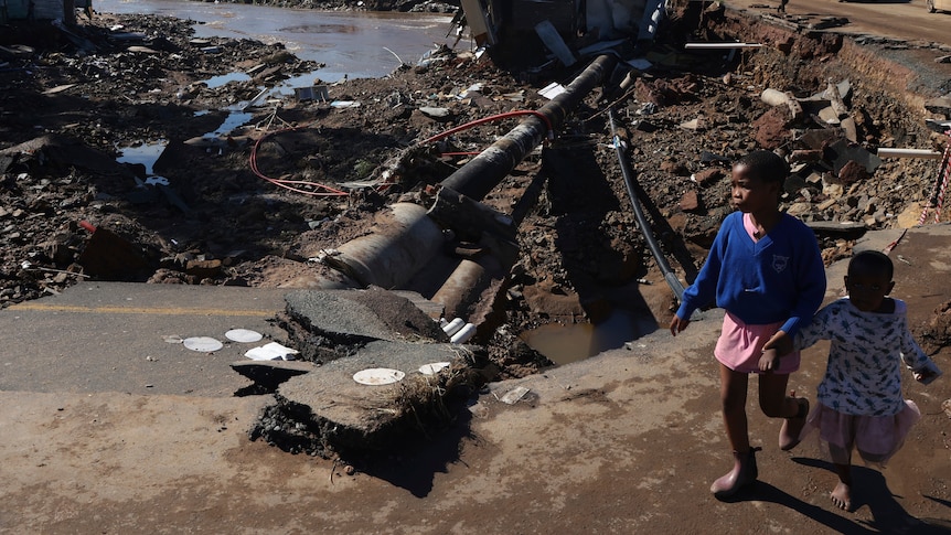 Two children holding hands walk near a damaged road, with pipes exposed, dirt and rubbish strewn.