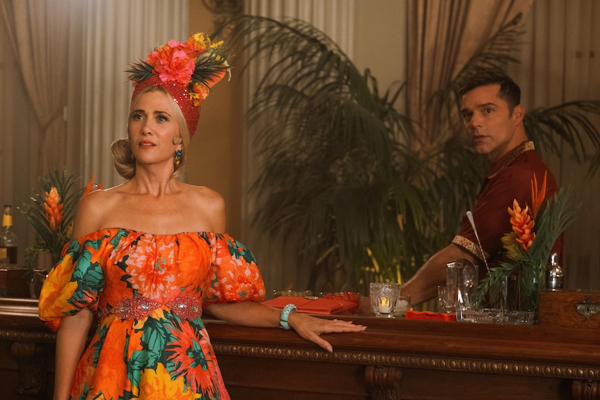 Kristen Wiig in a vibrant tropical print dress with matching fruit-filled headpiece. A bartender (Ricky Martin) is behind her.