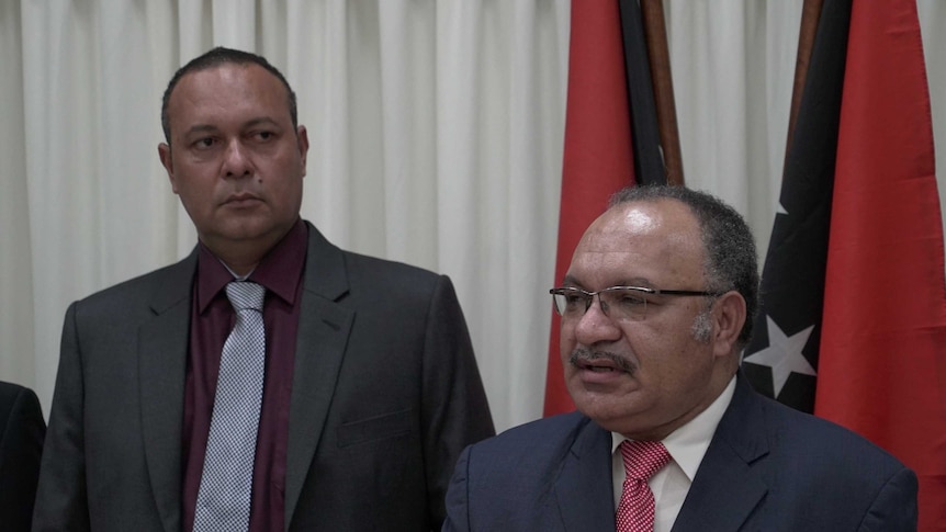 Two Papua New Guinean politicians speak to media with flags in the background.