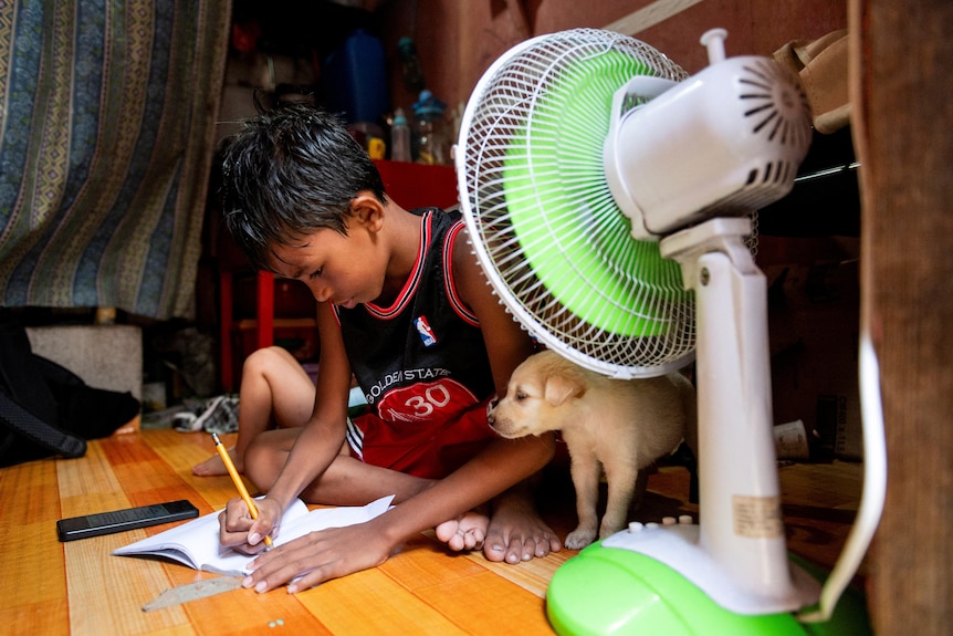A child works on their homework next to a puppy in front of fan.