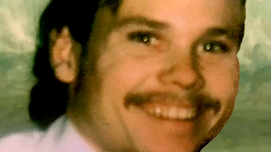 A picture of a young man smiling in a pink shirt with a moustache and long hair.