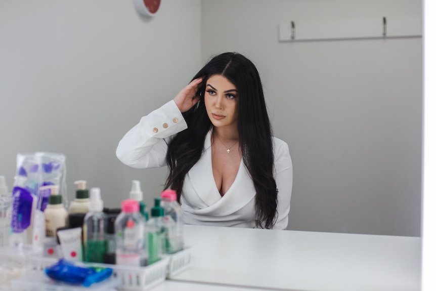 Natasha, with long brown hair and white jacket, looks into a mirror adjusting her hair.