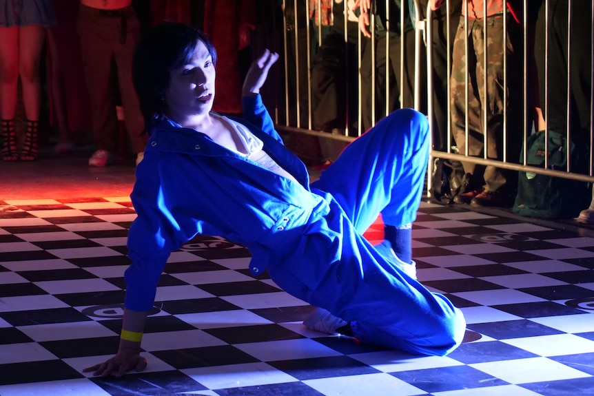An asian man wearing a blue jumpsuit poses on the floor. 