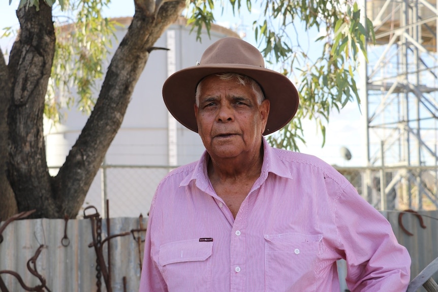 An elder stands under a gum tree with a brown, wide brimmed hat and pink shirt.