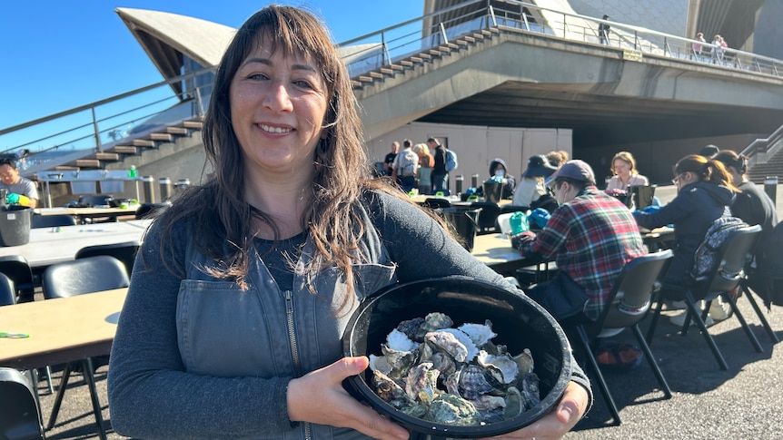 A woman holds a bucket of oyster shells in front of the Opera House.
