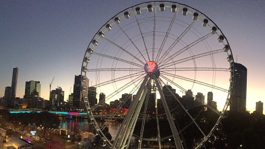 A ferris wheel called the brisbane wheel in front of the brisbane city skyline at sunrise.