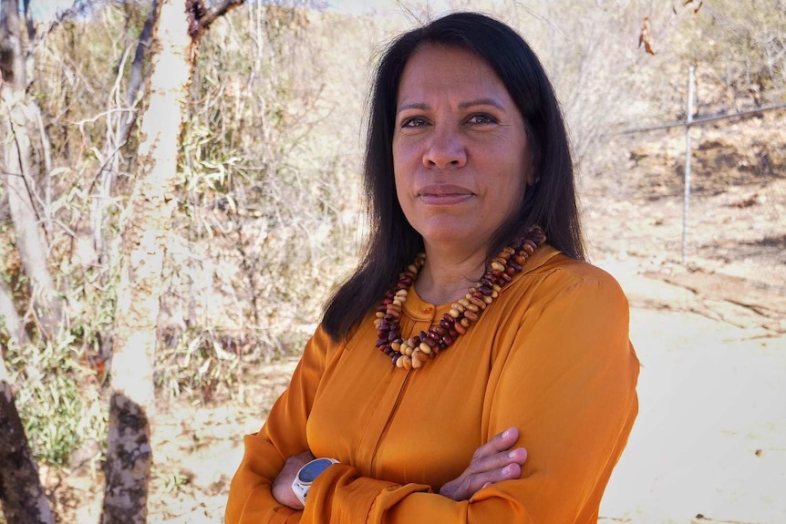 Dr Josie Douglas from the Central Land Council stands in front of trees and scrub in an orange dress.