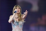 A close up of Taylor Swift singing into a microphone, reaching out a hand to the audience. She's wearing a sparkly leotard