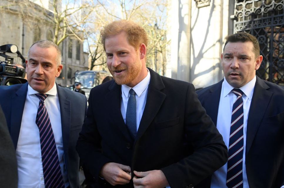 Prince Harry wears black suit and white shirt, smiles as he is surrounded by two men who look like bodyguards.