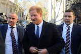 Prince Harry wears black suit and white shirt, smiles as he is surrounded by two men who look like bodyguards.