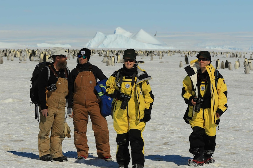 Four people stand on the ice in Antarctica. They are wearing protective clothing. There are penguins behind them.