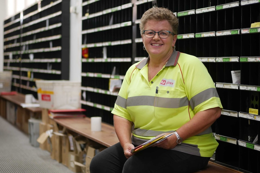 A woman in an Australia Post uniform sits on a bench while holding a handful of letters in a mail sorting centre.