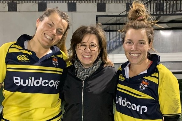 Jospehine Sukkar stands between two smiling rugby players wearing yellow and dark blue rugby kit