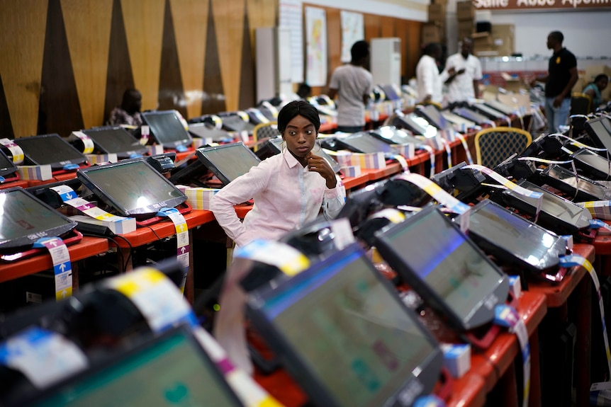 A woman in a pink shirt watches over three rows of touch screen computers printing election ballots.