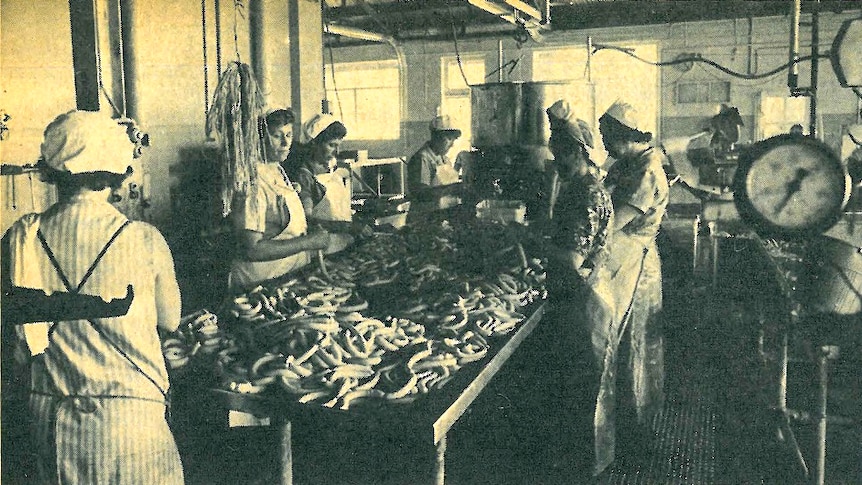 Women stand around a table of sausages inside a factory. The photo is black and white.