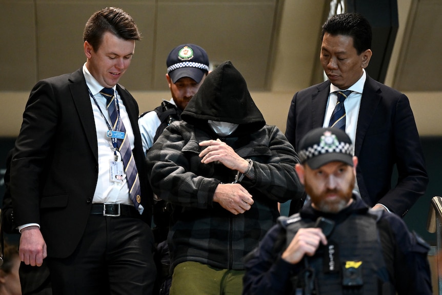 A man in handcuffs and wearing a hood and a face mask surrounded by police officers.
