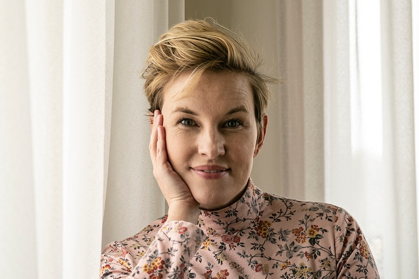 Actress with short blonde hair sits at table with white curtains behind, wearing floral top, and resting chin on hand.