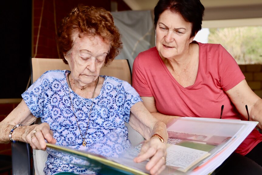 An old woman wearing a white and blue top looking through a photo album with her daughter wearing a pink top 