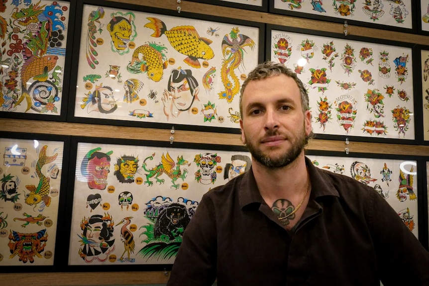 A young man wearing a black shirt, sits in front of a wall of tattoo designs. 