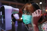A Thai woman in a face mask leans forward to have her temperature checked.