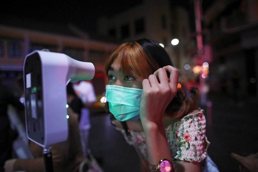 A Thai woman in a face mask leans forward to have her temperature checked.