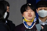 Korean man wearing neck brace speaks in front of microphone, surrounding by five people wearing masks.