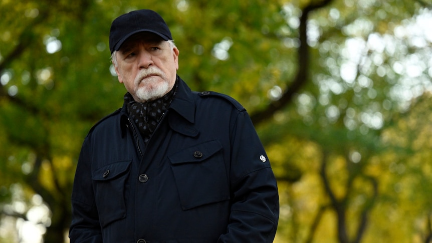 Logan stands in a black jacket and cap amongst trees and looks off seriously into the distance.