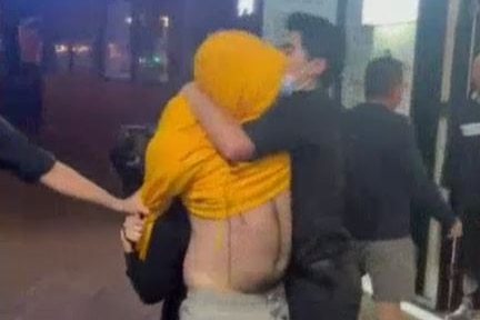 A person with a yellow hooded jumper over their face and stomach exposed is held by people wearing all black
