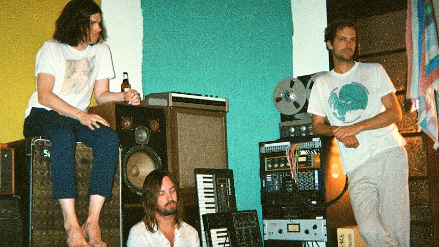 Photo of Tame Impala in studio, Kevin Parker seated on the floor, Jay leaning against gear, Dom sitting on a speaker stack