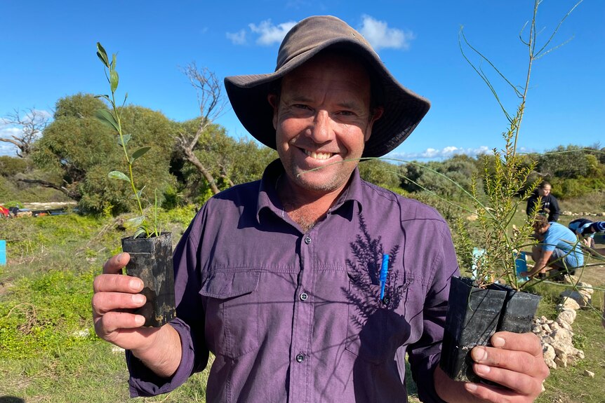 Man in hat holding up seedling plants in either hand with a smile looking at the camera, tree planting in bush in background
