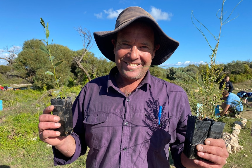 A man in a hat holds up seedlings in either hand with a smile, looking at the camera. Students plant trees behind him.