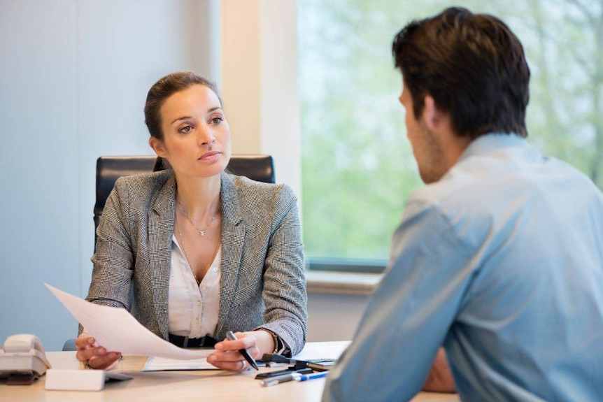 In an office room, a woman in jacket holds paper and pen while looking man in shirt, seen from the back.