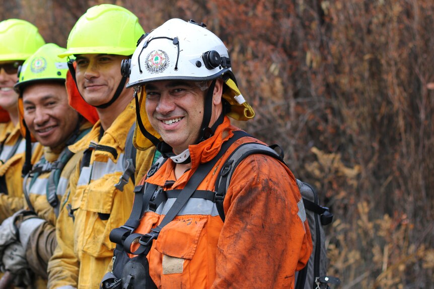 A group of smiling firefighters standing in front of burnt out bush