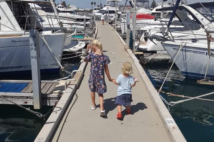 A young girl holds the hand of a toddler as they walk along a jetty.