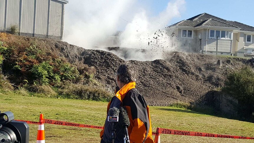 A man standing in front of a mound of dirt with a puff of steam and mud erupting from the pit.